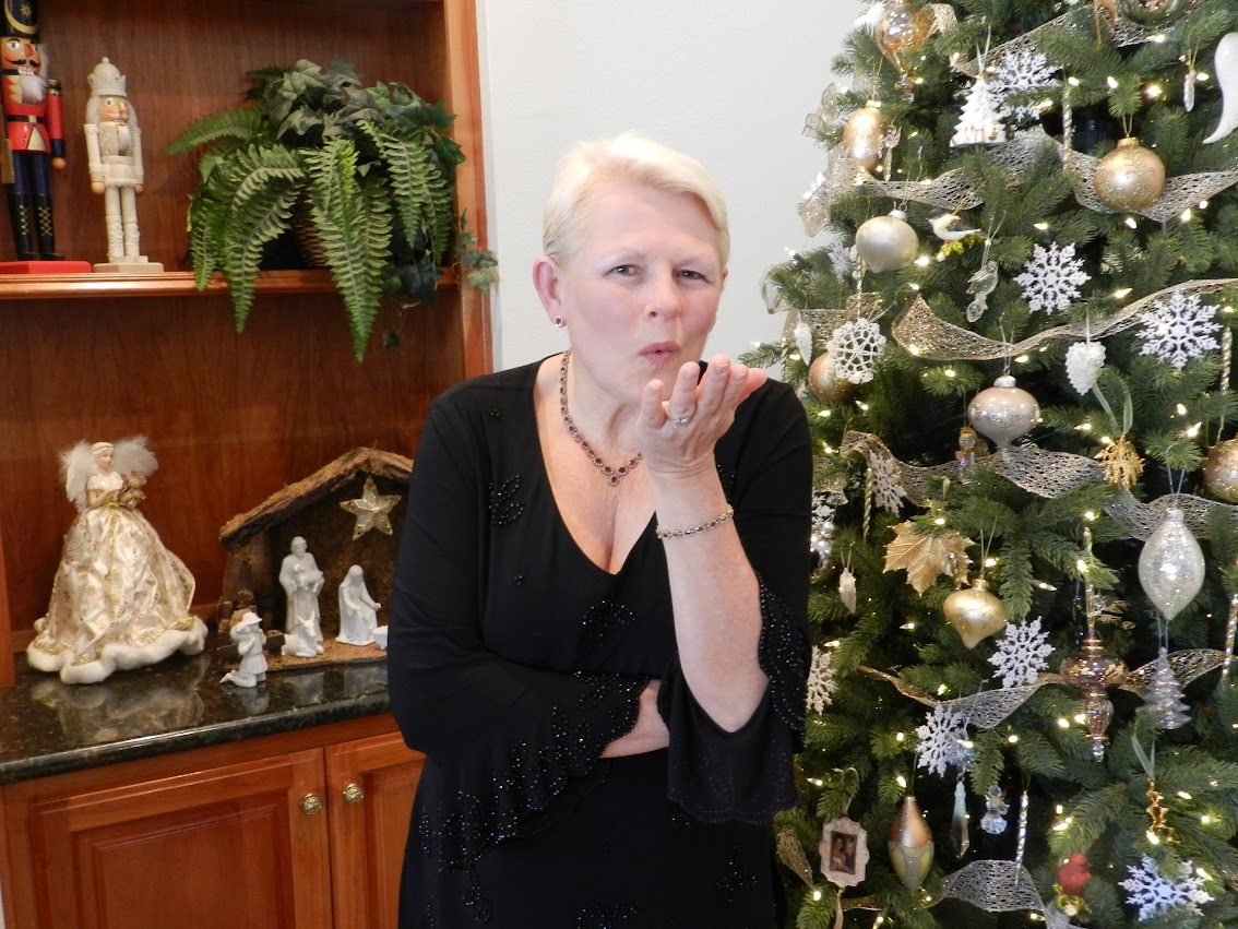A smiling woman in a black top blowing a kiss beside a Christmas tree decorated in gold and silver