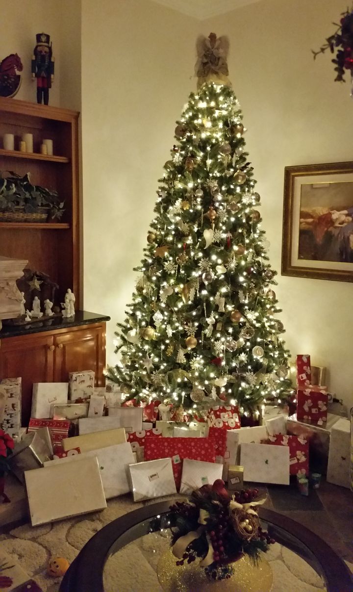 A tall Christmas tree glowing with white lights in a living room corner, with a pile of wrapped gifts underneath