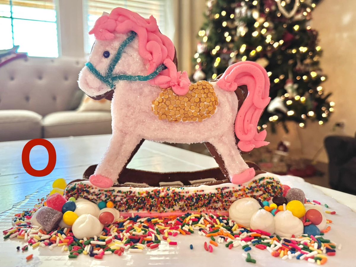 A gingerbread rocking horse with a pink frosting mane and tail, gold sprinkle saddle and a candy base, in front of a Christmas tree